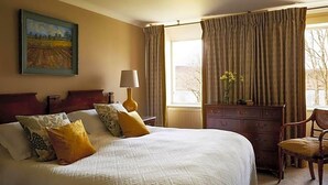 A neatly made bed with a headboard, a bedside table with a lamp, a dresser, and a chair. There is a window with curtains, and a painting on the wall. - Kinloch Lodge (Sleat)