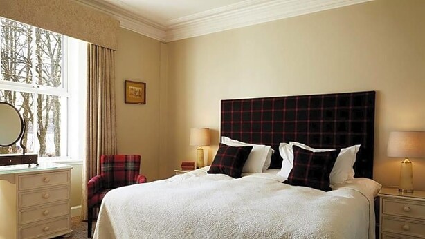 A hotel room with a large bed, a dresser, a chair, and a nightstand with a lamp. - Kinloch Lodge (Sleat)