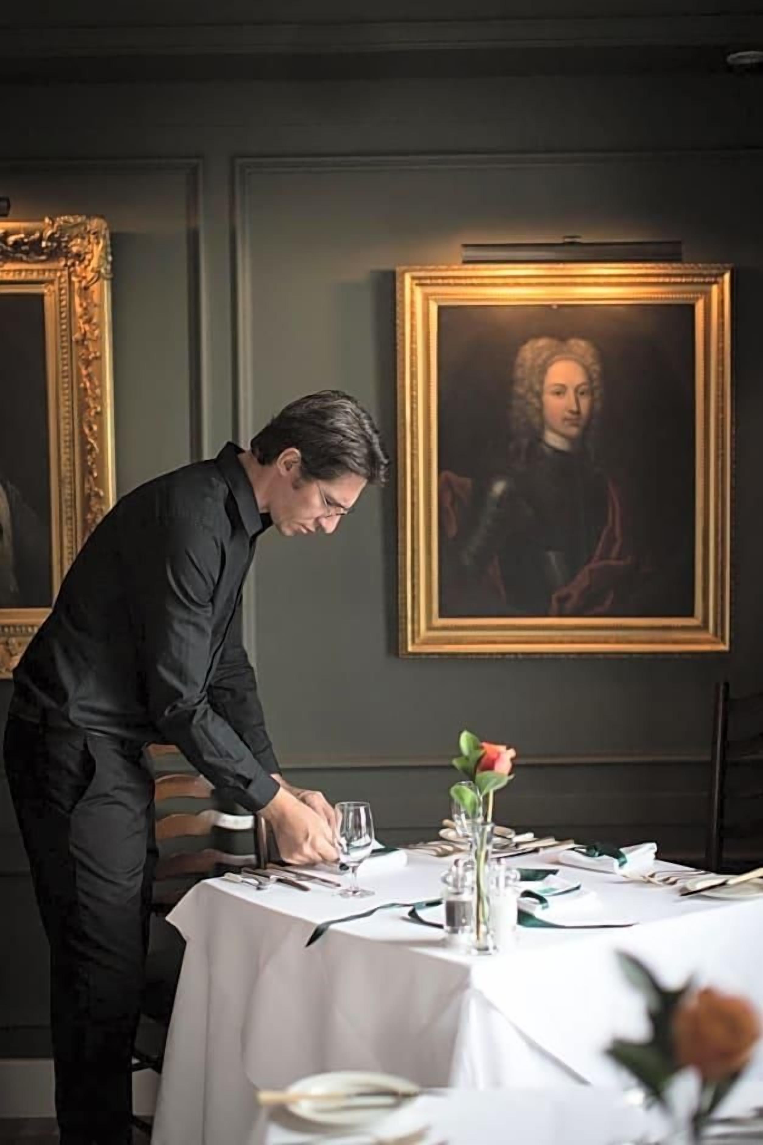 A person setting a table with a white tablecloth, a vase with a single flower, and a framed painting on the wall.