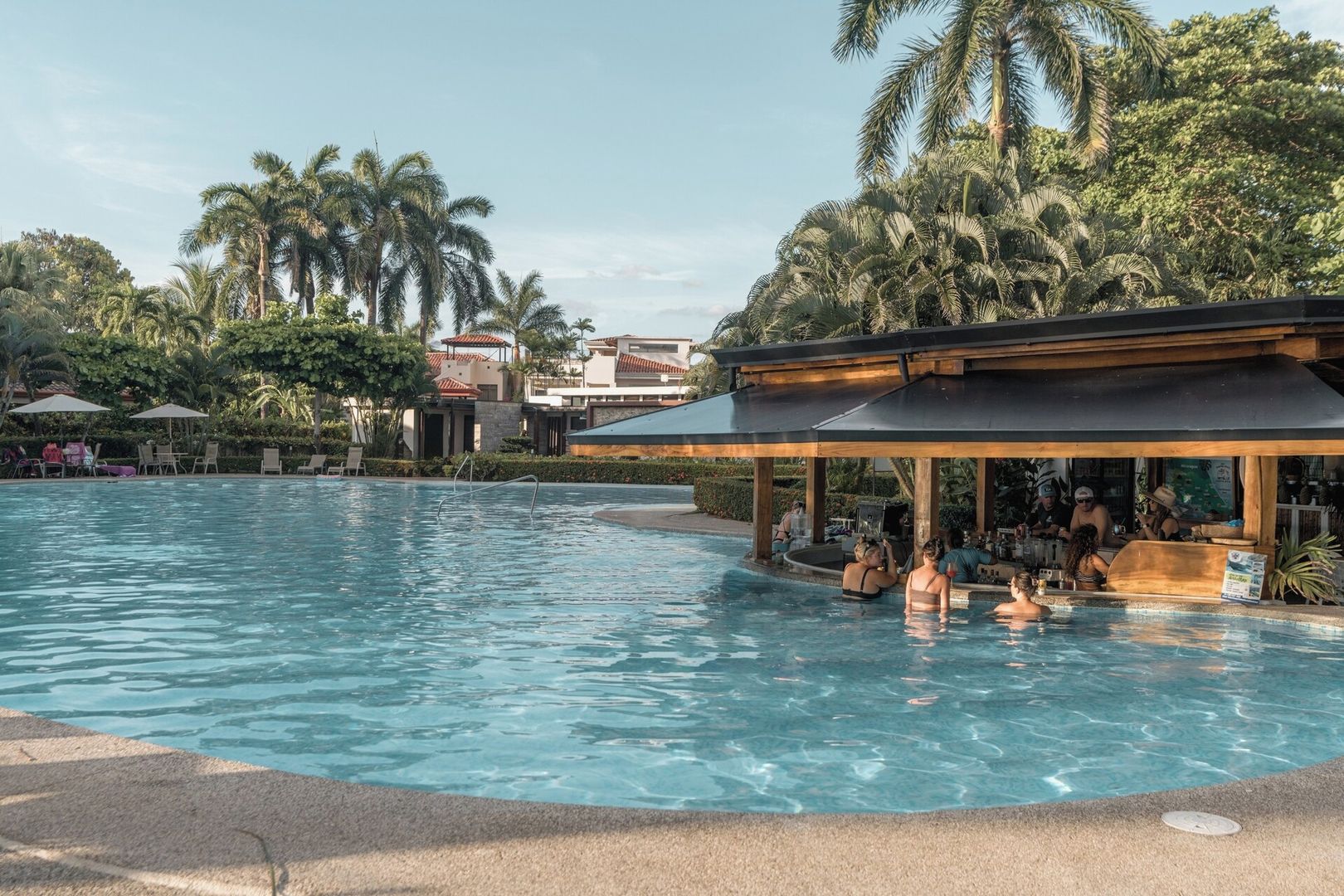 Una piscina con sillones de descanso y sombrillas, rodeada de palmeras y una zona de bar techada.