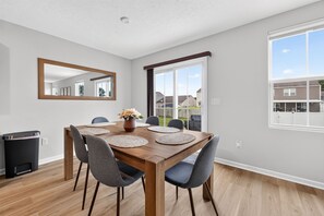 Een eetruimte met een houten tafel, zes stoelen, een spiegel en een raam met uitzicht op huizen.