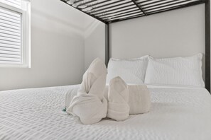 Een net opgemaakt bed met witte lakens en kussens, een raam met zonwering en een metalen hoofdbord.