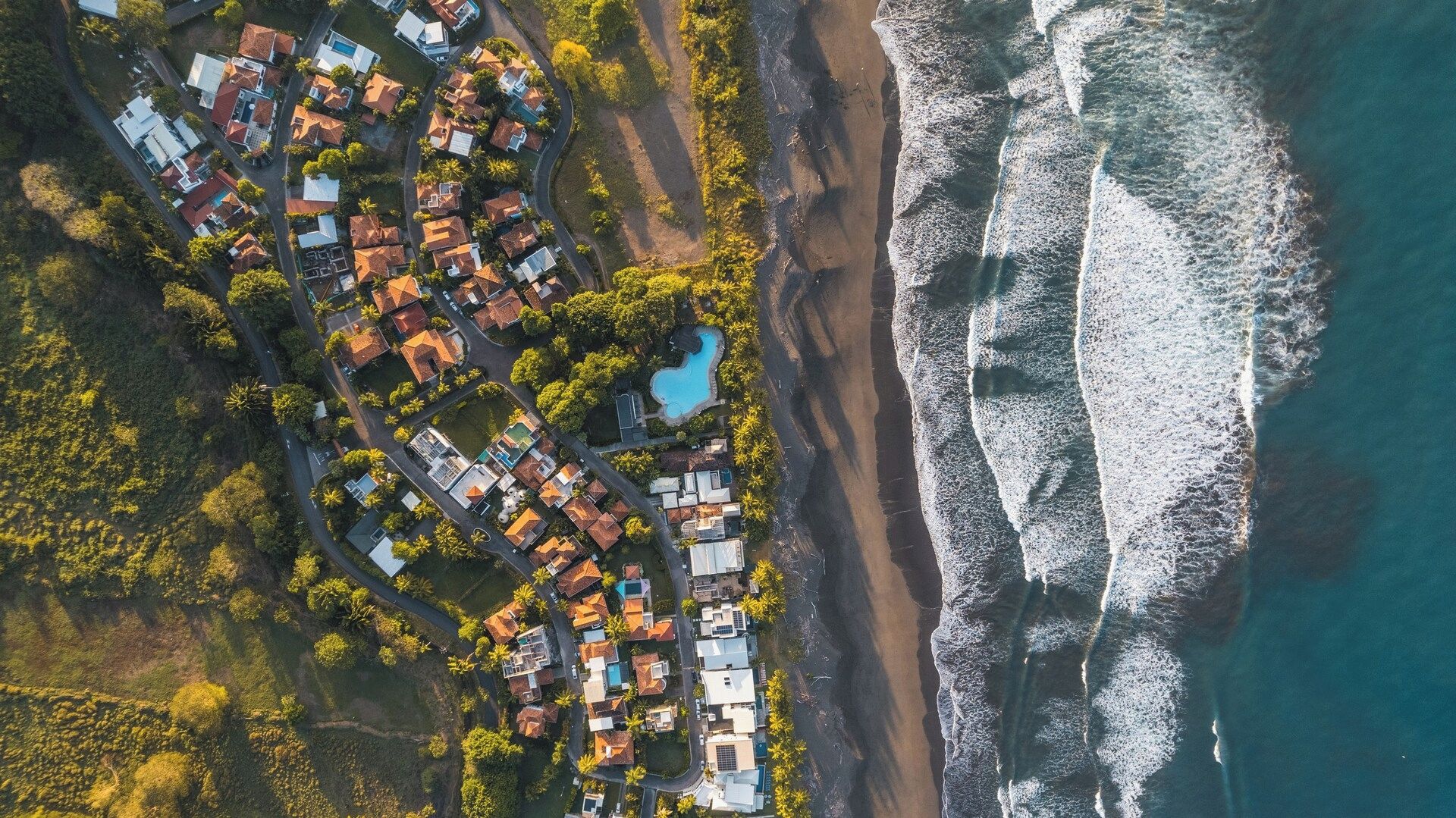 An aerial view of a coastal residential area with houses, a swimming pool, and a beach.