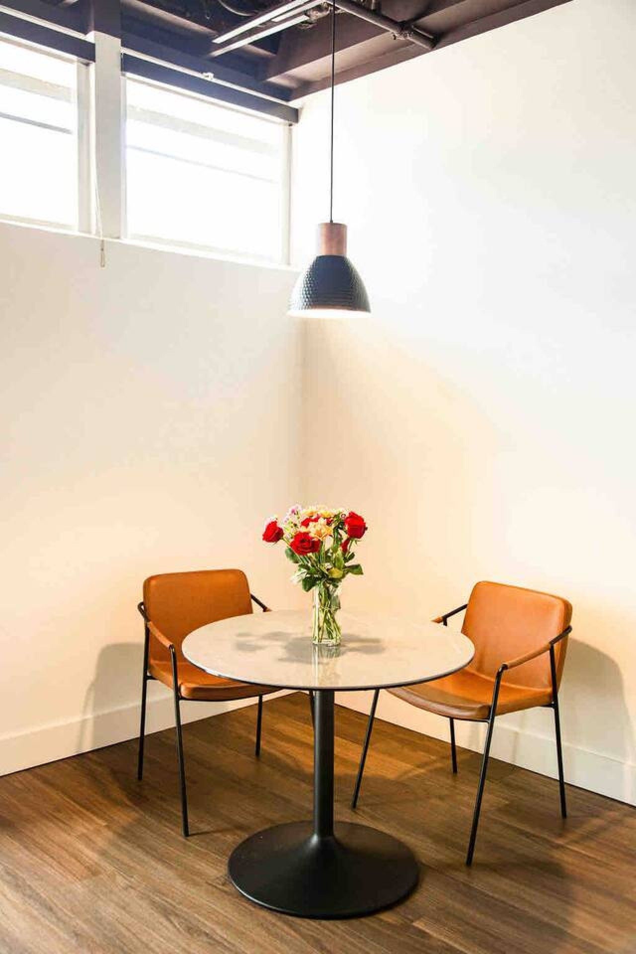 A round table with a black base, two orange chairs, and a vase with flowers.