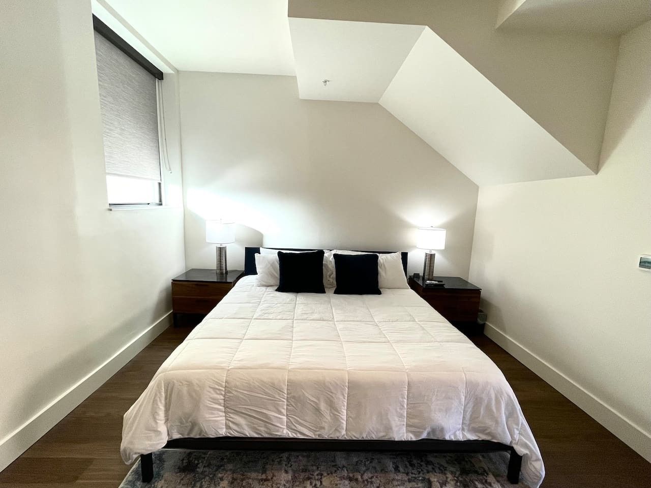Quarto com cama, duas mesas de cabeceira, janela com persianas e teto inclinado.