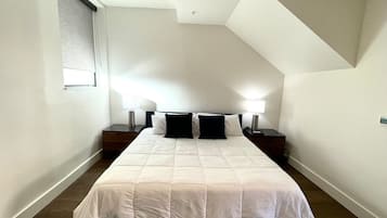 Quarto com cama, duas mesas de cabeceira, janela com persianas e teto inclinado.