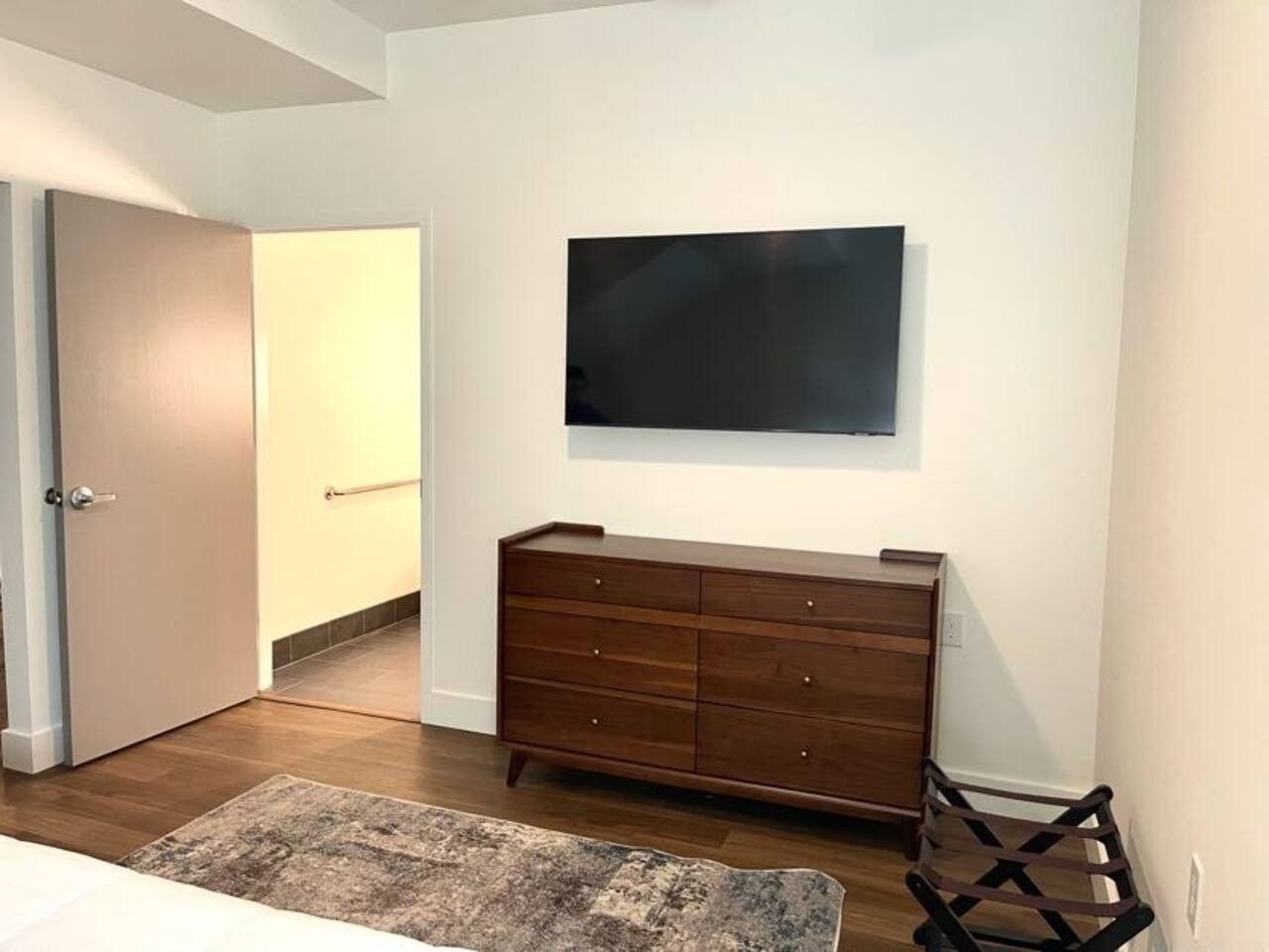 A room with a wooden dresser, a flat-screen TV mounted on the wall, a door, and a rug on the floor.