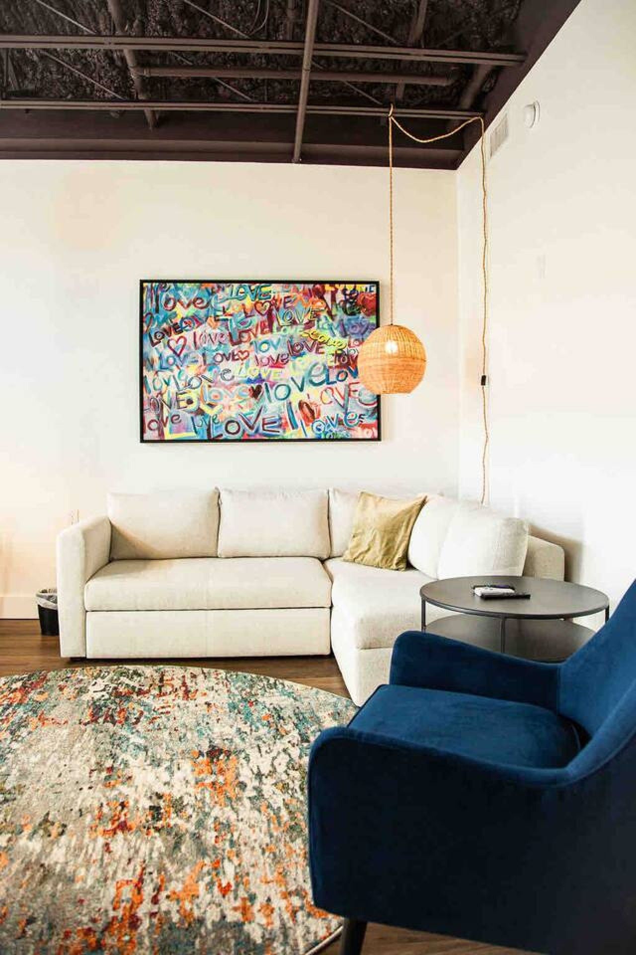Un salon moderne avec un canapé, un fauteuil bleu, une table basse ronde, un tapis coloré et une peinture abstraite aux couleurs vives accrochée au mur.