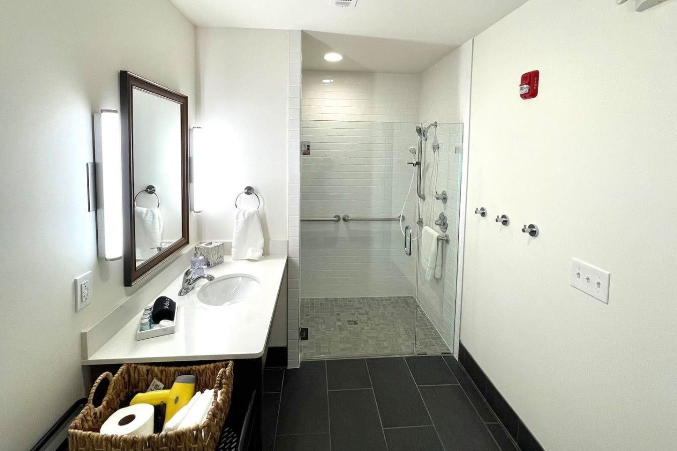 A modern bathroom with a dual sink, a large mirror, a shower area, and a basket of toiletries.