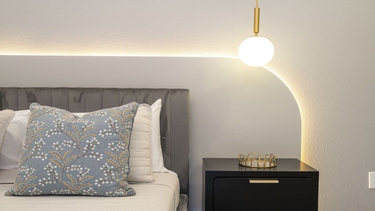 Cama bem arrumada com travesseiro floral, mesa de cabeceira com abajur e um objeto decorativo sobre a mesa.