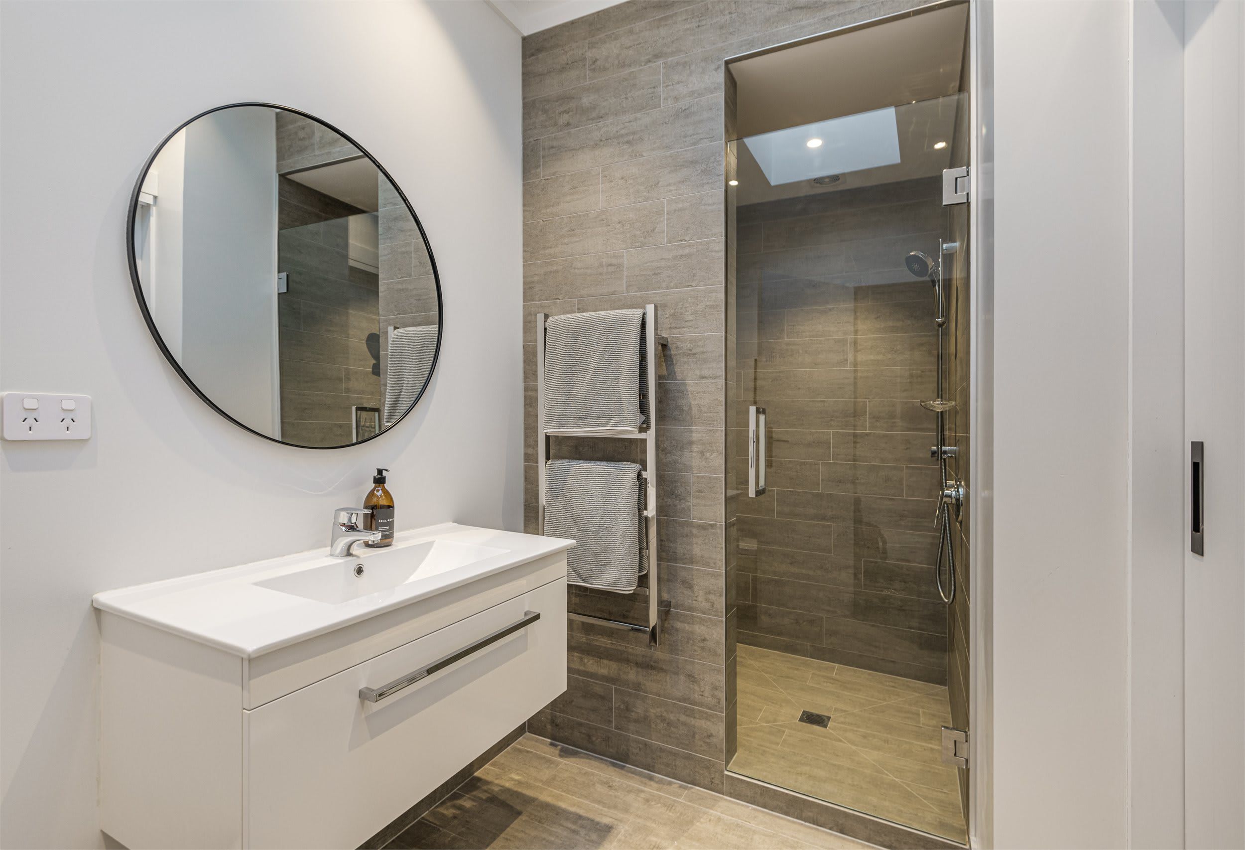 A modern bathroom with a white vanity, a round mirror, a glass shower enclosure, and a towel rack.