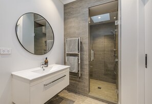 Un baño moderno con un tocador blanco, un espejo redondo, cabina de ducha con mampara de vidrio y un perchero para toallas.