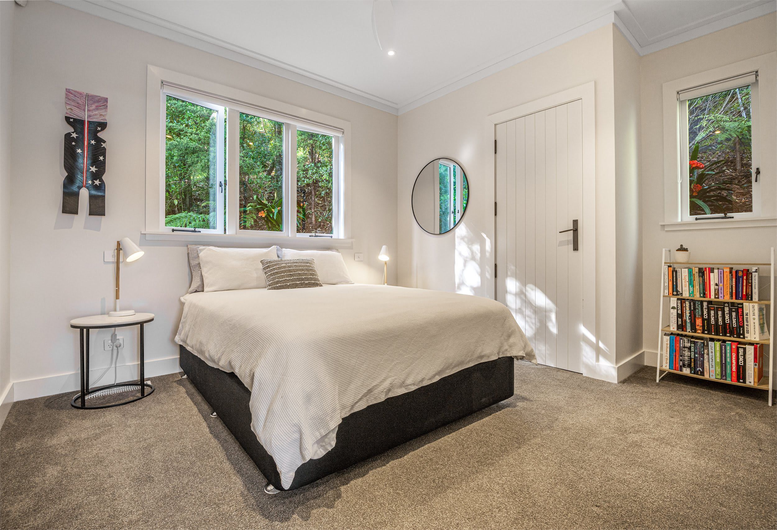A bedroom with a bed, bedside tables, a bookshelf, and large windows.