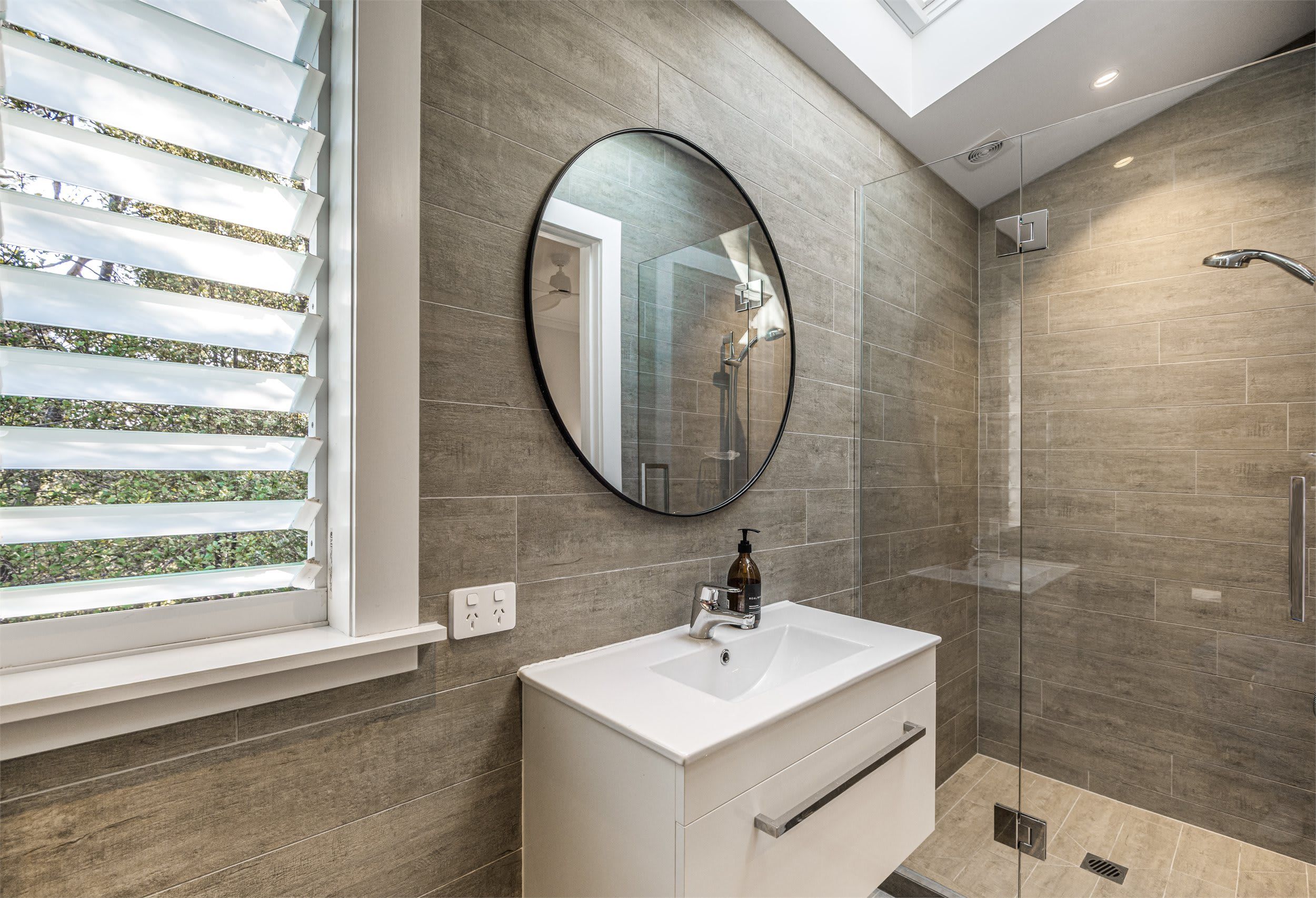 A modern bathroom with a large mirror, a glass shower enclosure, and a window with blinds.
