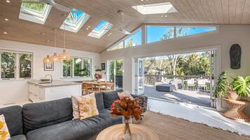 A modern living room with a grey sofa, a dining area, and a view of the outdoors through large windows.