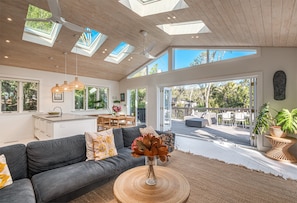A modern living room with a grey sofa, a dining area, and a view of the outdoors through large windows.