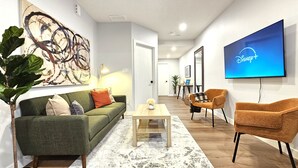 Una sala de estar moderna con un sofá verde, una mesa de centro de madera, un cuadro abstracto de gran tamaño, un televisor mostrando el logotipo de Disney+ y una planta en maceta.