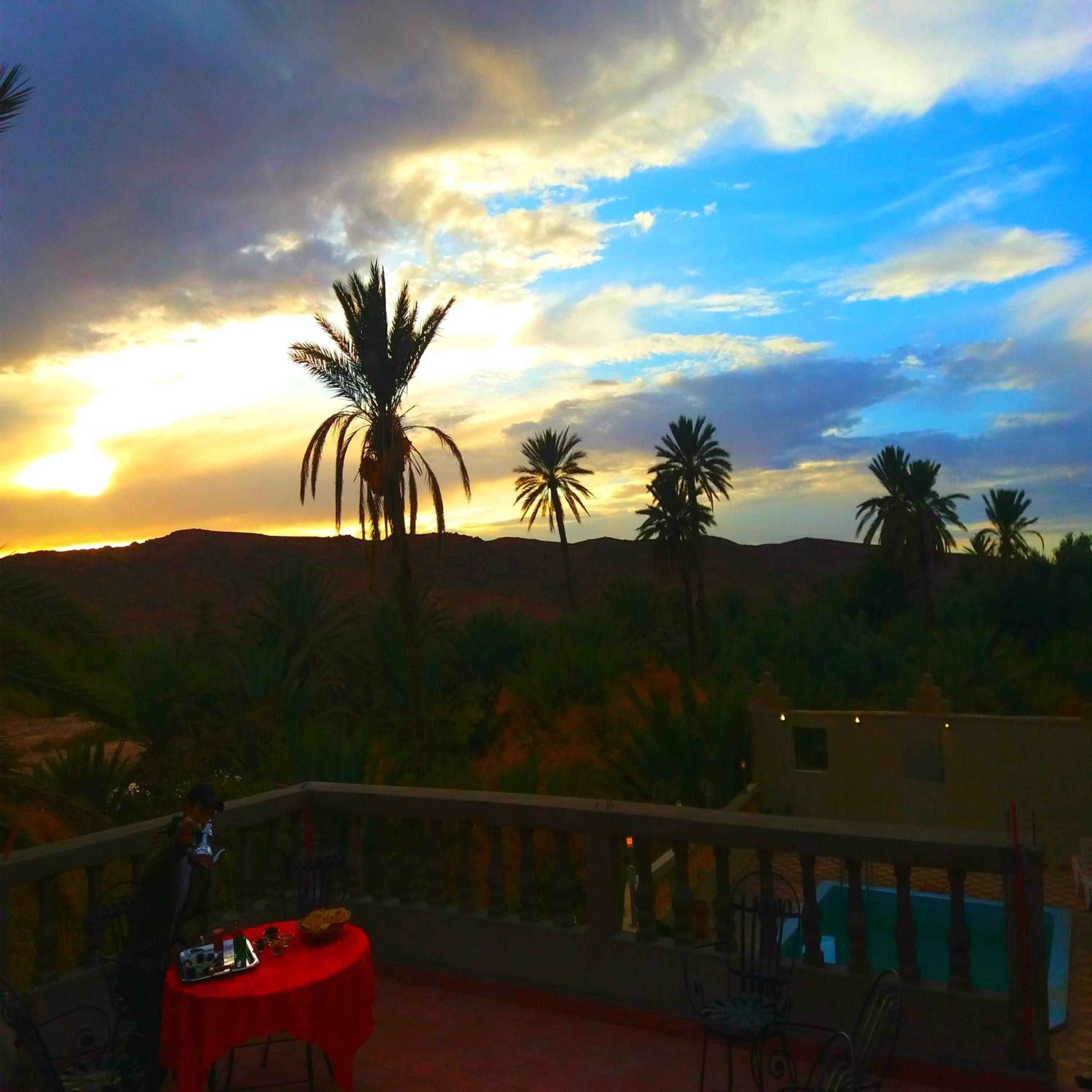 A terrace with a table and chairs, overlooking a pool and palm trees against a sunset sky.