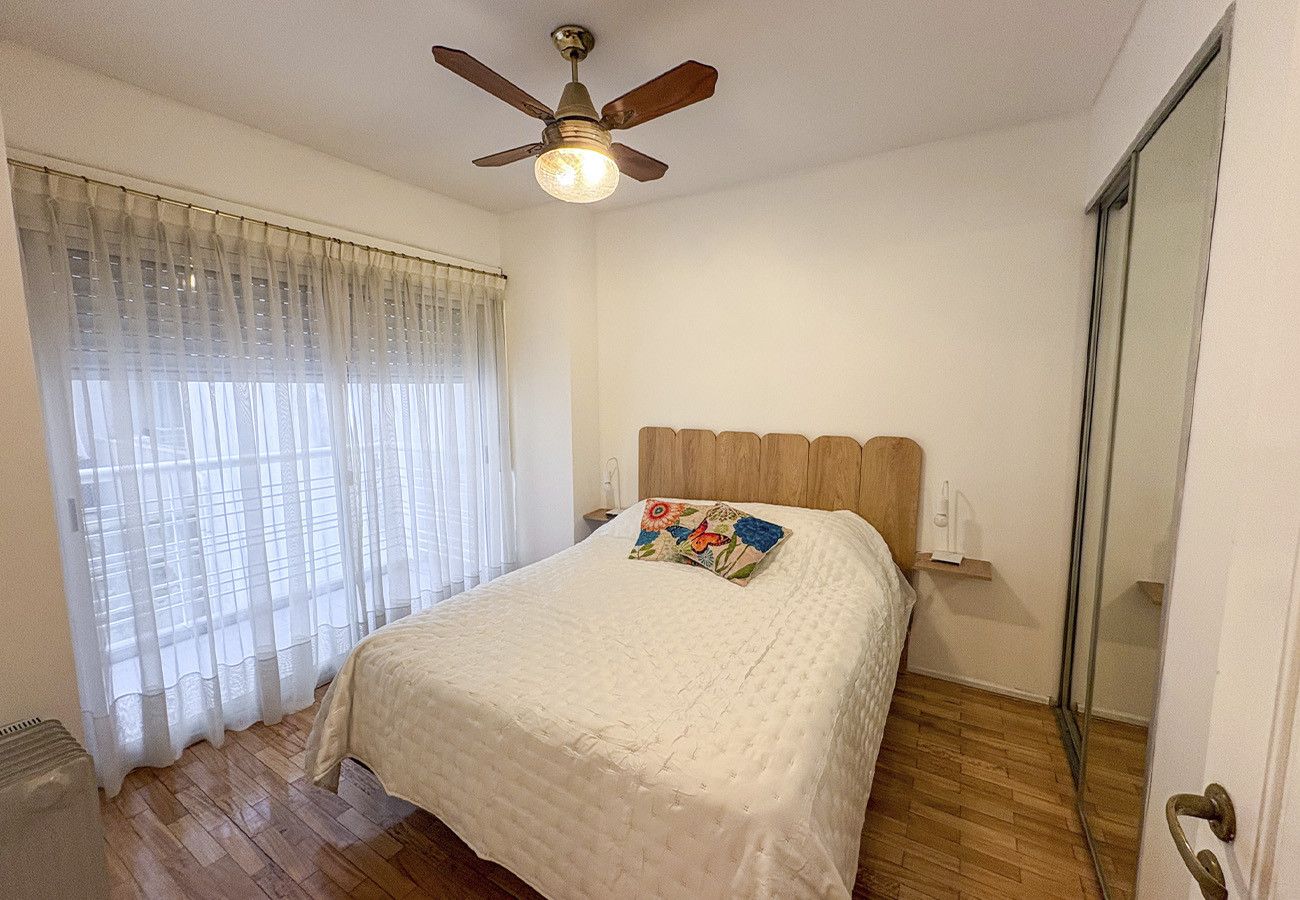A bedroom with a bed, a ceiling fan, a window with sheer curtains, and a wooden floor.