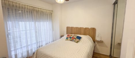 A bedroom with a bed, a ceiling fan, a window with sheer curtains, and a wooden floor.