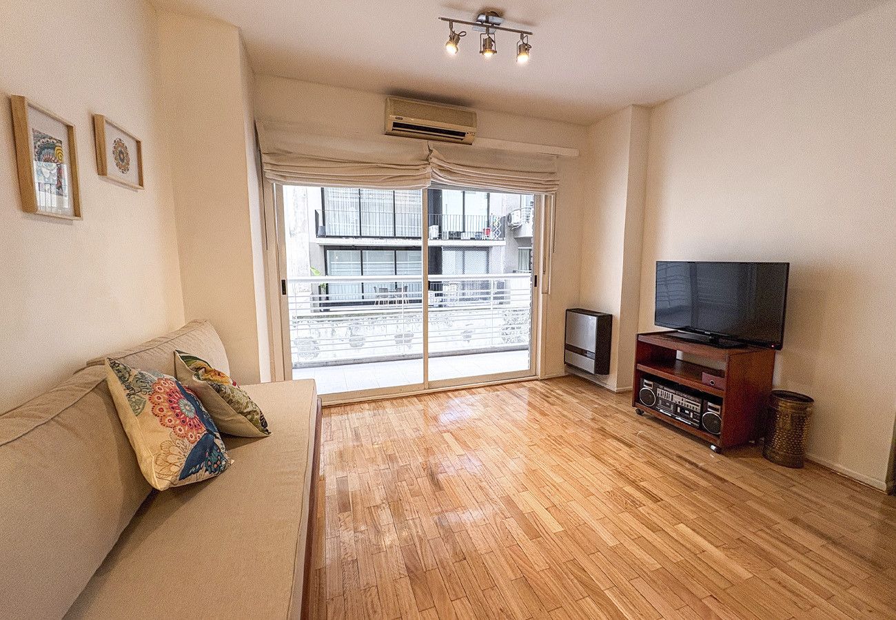 Uma sala de estar com sofá, televisão e uma porta de vidro deslizante que dá para uma varanda.
