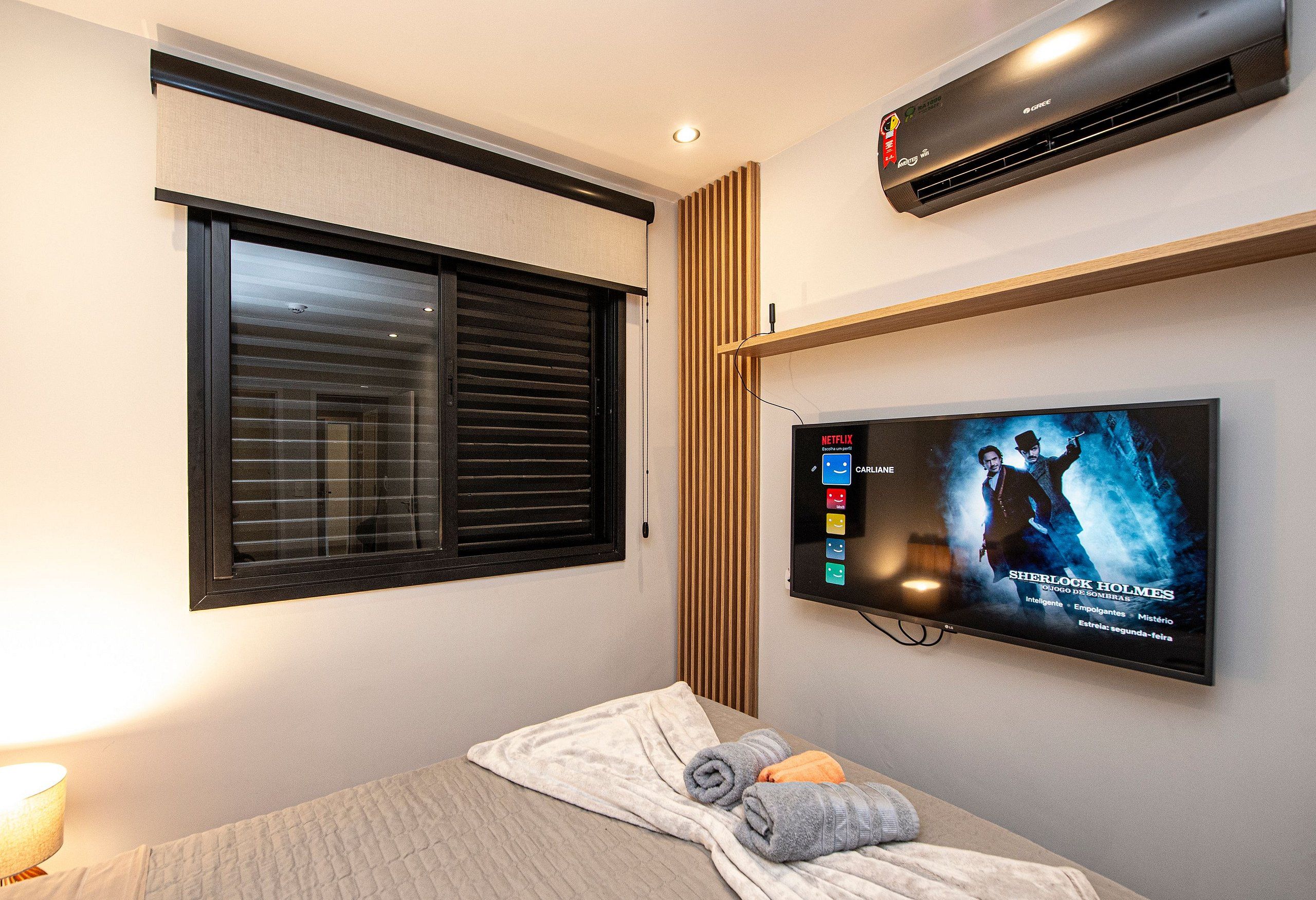Una habitación de hotel con cama, televisión mostrando un programa, ventana con persianas y aire acondicionado.