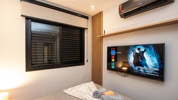 Una habitación de hotel con cama, televisión mostrando un programa, ventana con persianas y aire acondicionado.