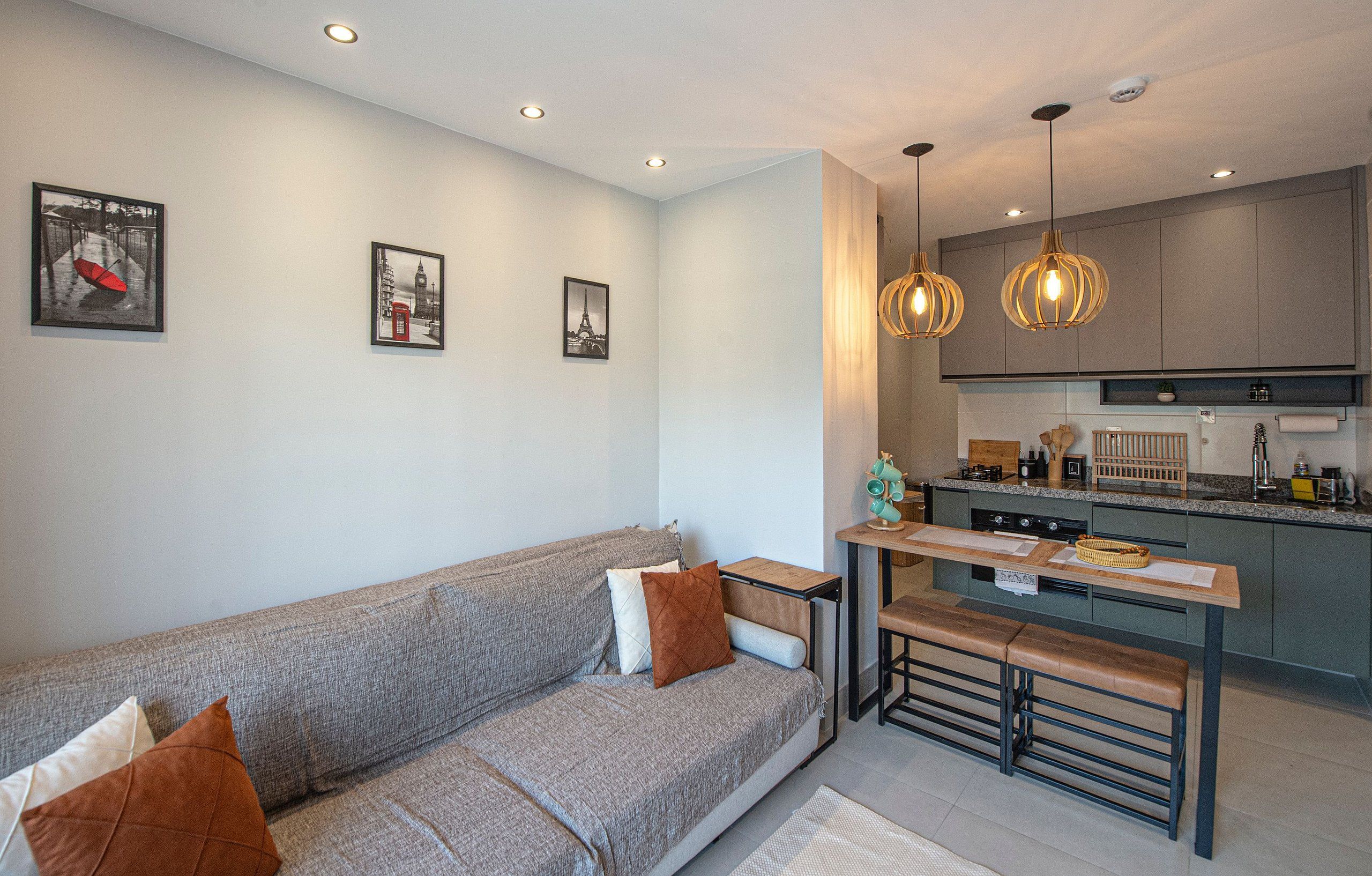 A modern living room with a grey sofa, wooden bench, and a kitchen area with grey cabinets and pendant lighting.