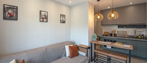 Ein modernes Wohnzimmer mit grauem Sofa, einer Holzbank und einer Küchenzeile mit grauen Schränken und Pendelleuchten.