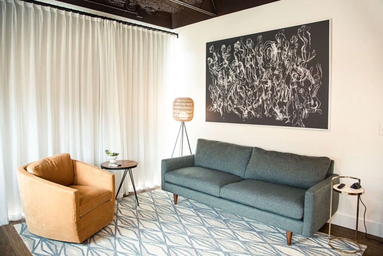 Uma sala de estar com sofá, poltrona, tapete estampado, luminária de chão e uma grande pintura abstrata na parede.
