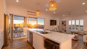 A modern kitchen with a large island, sliding glass doors leading to a balcony, and a dining area with a view. - Dora's Place | Pet Friendly | Sunset Views (Agnes Water)