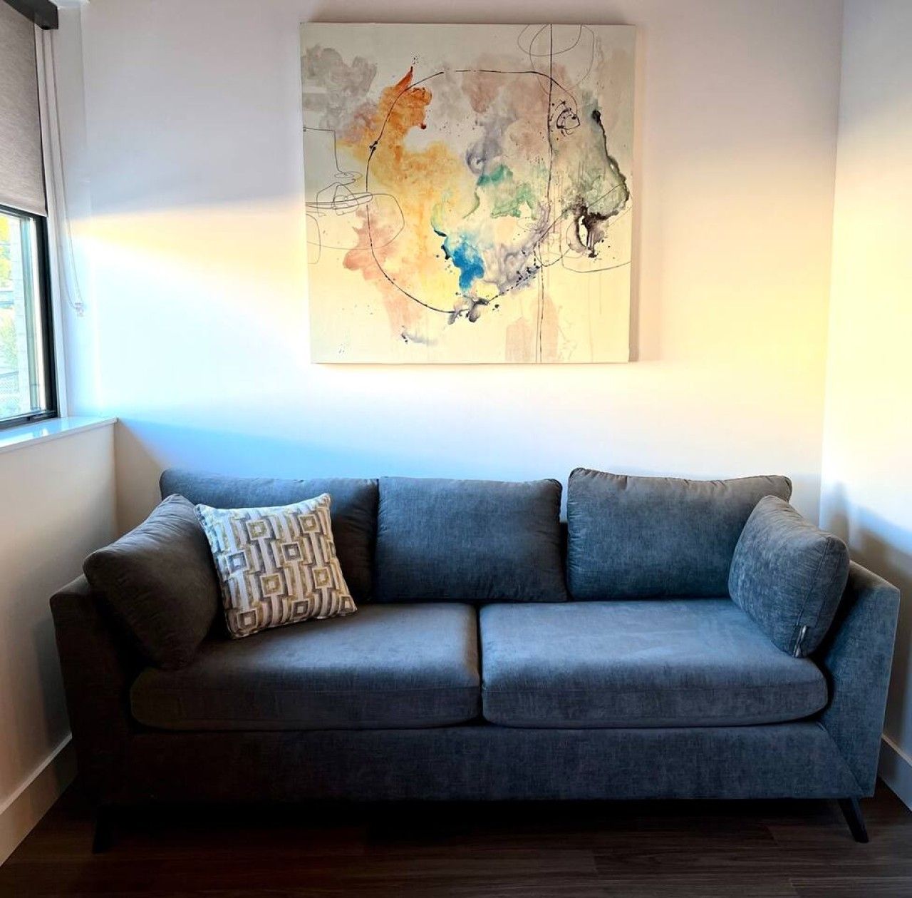 Um sofá cinza com uma almofada estampada em frente a uma parede com um quadro abstrato colorido.