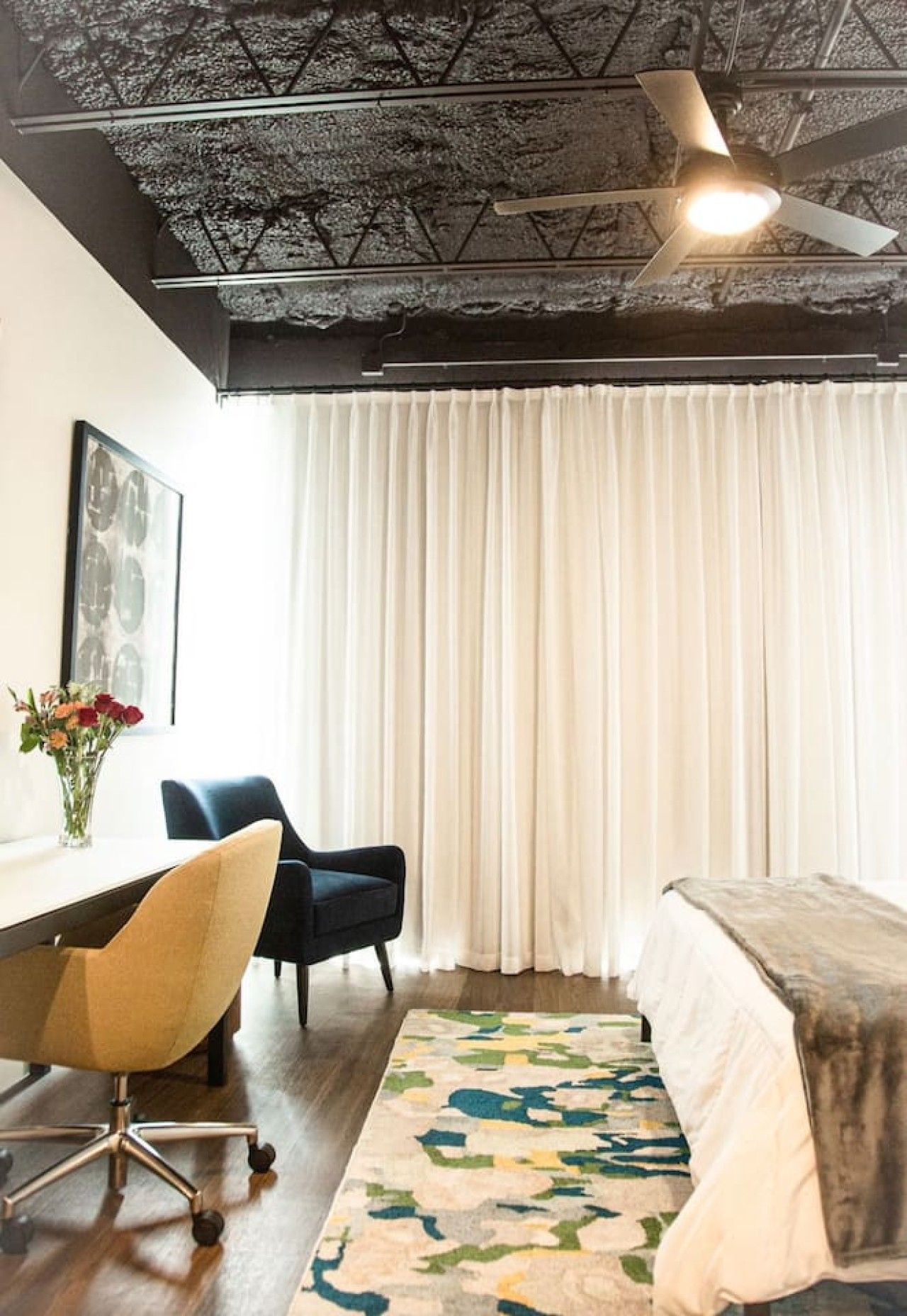 Quarto de hotel com cama, escrivaninha, cadeira, ventilador de teto e janela com cortinas.