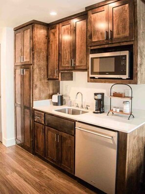 A kitchen with wooden cabinets, a stainless steel dishwasher, a microwave, and a coffee maker. - The Lofts Suite #21 `The Judge` King Bed & Balcony (Wetumpka)