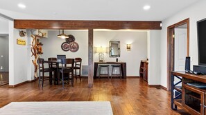 A spacious living area with a wooden floor, a dining table with chairs, a TV stand, and a wall-mounted clock. - Poolside Pines Reunion House by Avantstay Private Pool + Hot Tub + Gym Mins to the Lake (Twin Peaks)