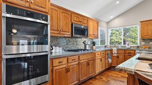 A modern kitchen with stainless steel appliances, wooden cabinetry, and a stone backsplash. - Poolside Pines Reunion House by Avantstay Private Pool + Hot Tub + Gym Mins to the Lake (Twin Peaks)