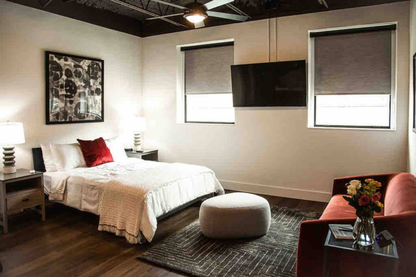 Un dormitorio moderno con cama, mesitas de noche, almohada roja, otomana blanca, sofá rojo, televisor montado en la pared y un ventanal grande.