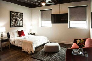 Un dormitorio moderno con cama, mesitas de noche, almohada roja, otomana blanca, sofá rojo, televisor montado en la pared y un ventanal grande.