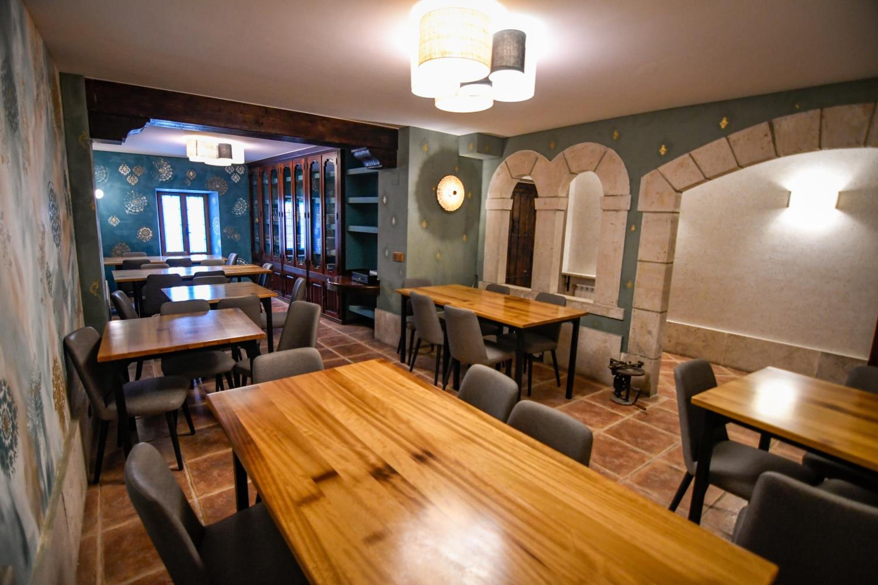 Interior de restaurante con mesas y sillas de madera, pared azul con motivos decorativos y un arco de piedra.
