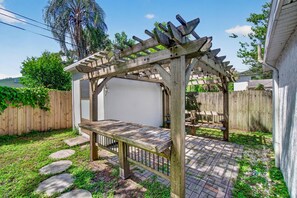 Een houten pergola met een bankje, een terras van bakstenen en een houten omheining in een achtertuin.