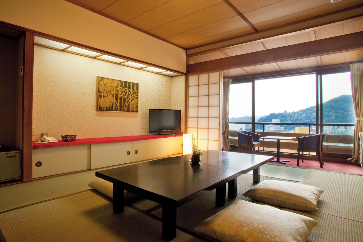 A traditional Japanese room with tatami flooring, a low table, a television, and a view of a mountainous landscape.