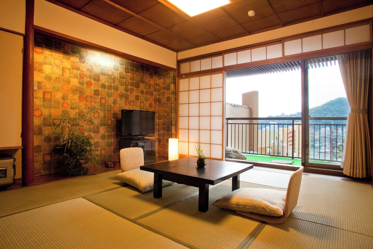 A traditional Japanese room with tatami flooring, a low table, a sofa, a TV, and a view of a cityscape through large windows.