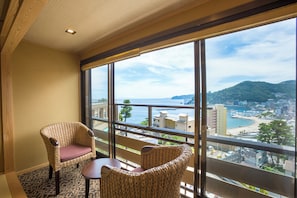 A balcony with wicker chairs and a table, overlooking a coastal cityscape with buildings and a beach. - atamionsen yuyadoichibanchi (Atami)