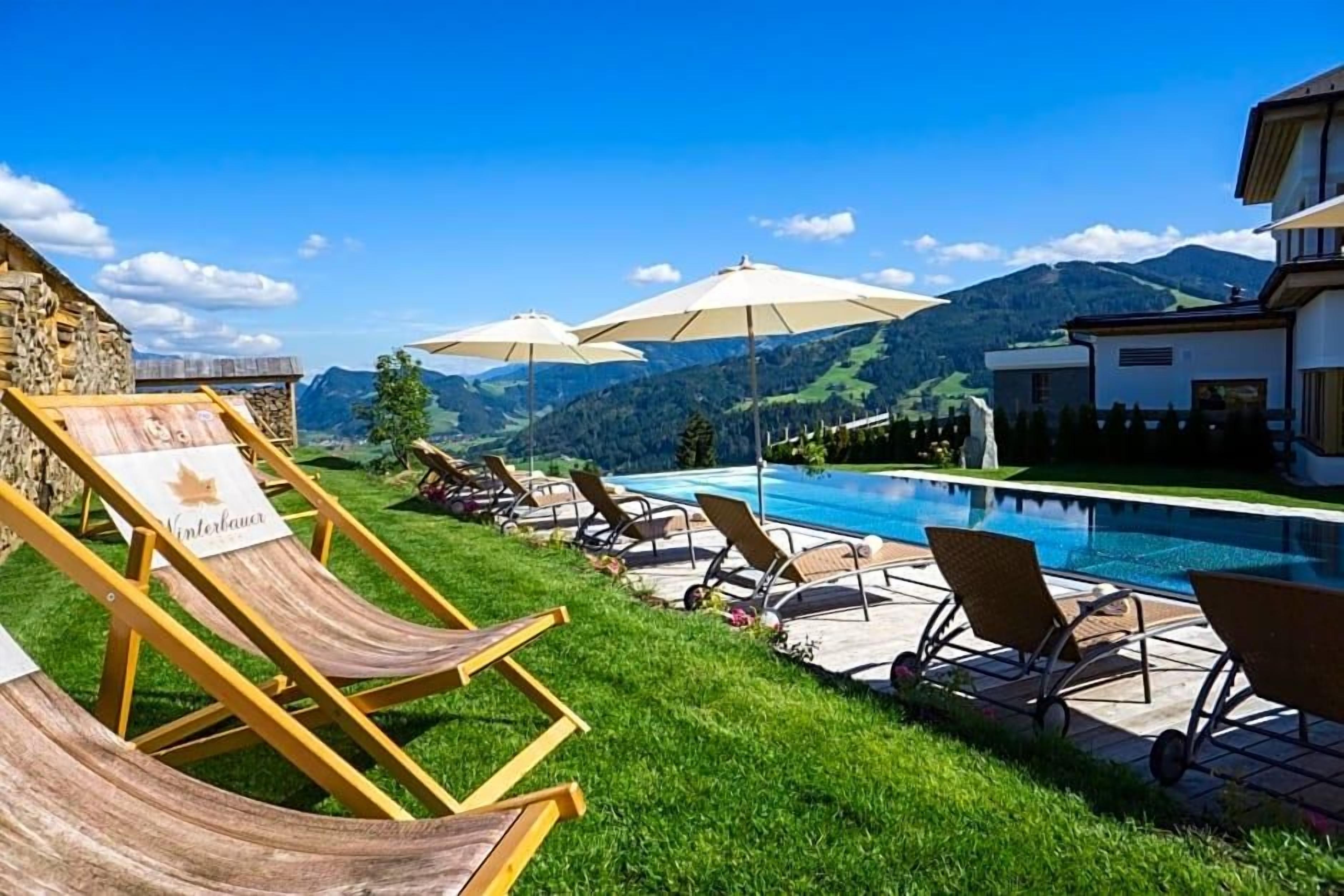 Ein Poolbereich mit Liegestühlen und Sonnenschirmen, eine Steinmauer und Blick auf Berge.