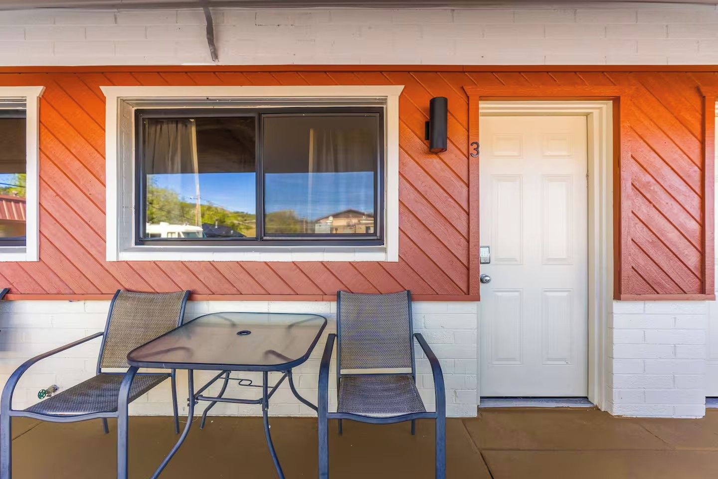A covered patio with a table and two chairs, a white door, and a window with a view of houses.