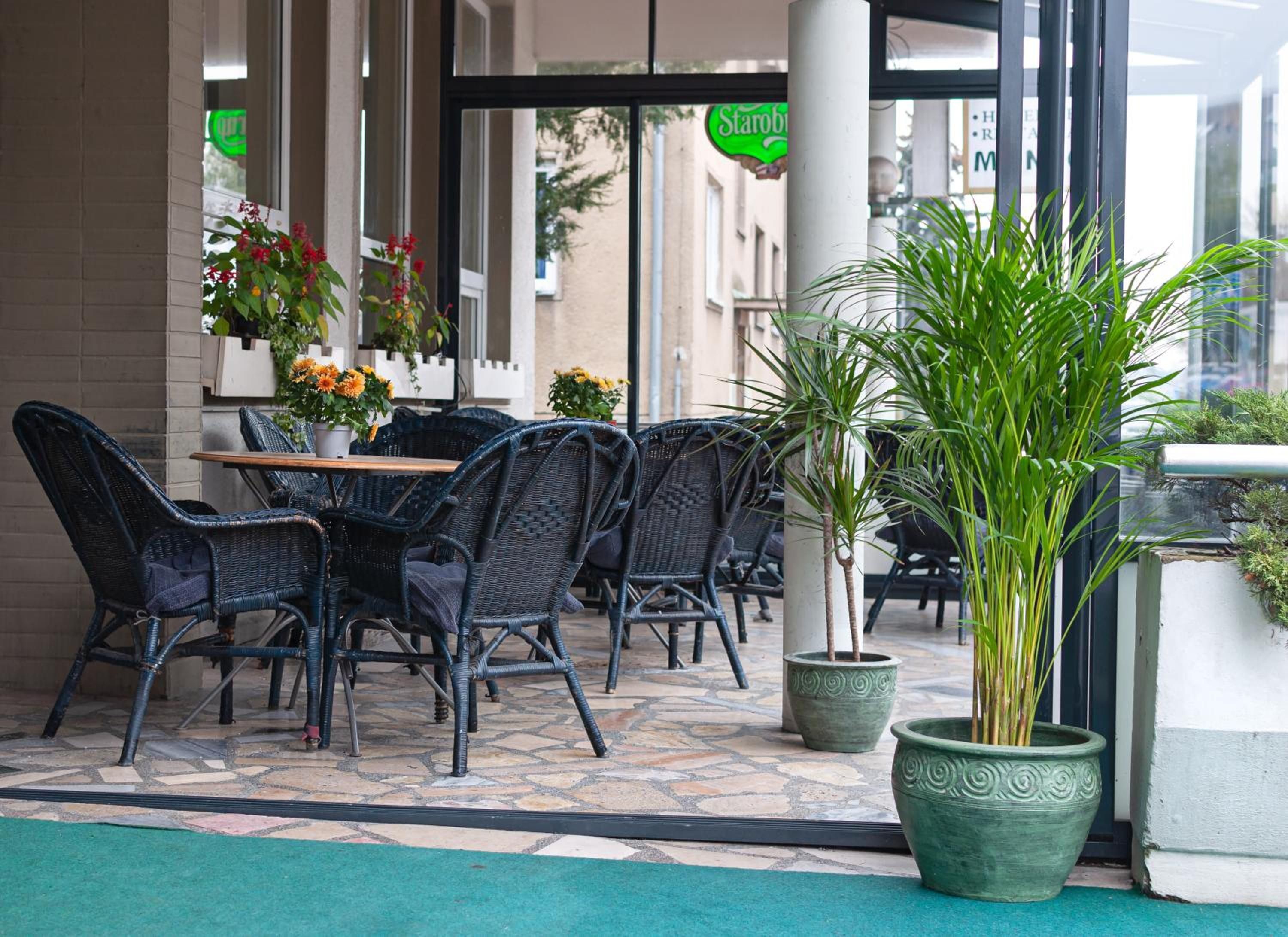 An outdoor seating area with wicker chairs and tables, potted plants, and a building entrance with a sign.