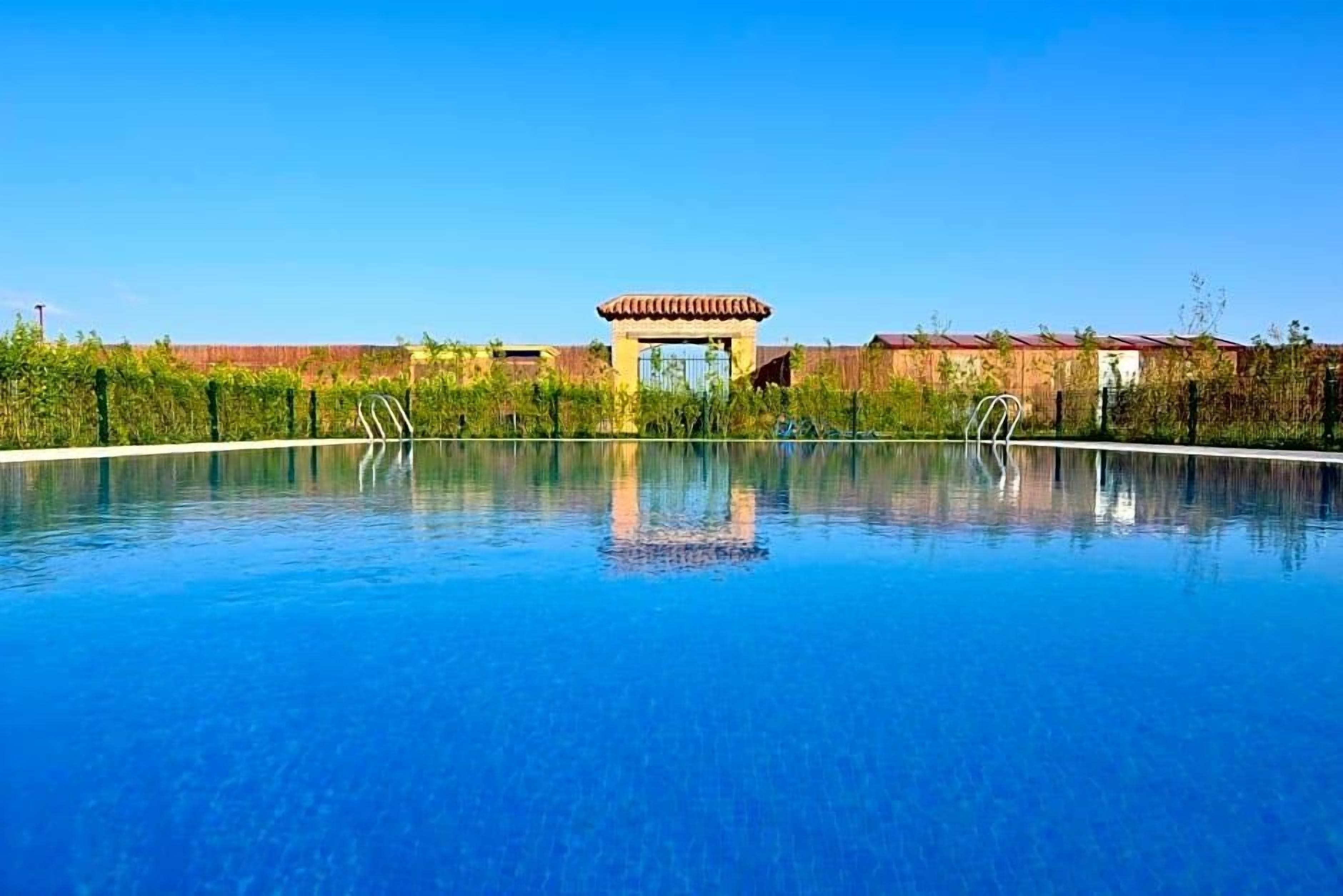 A clear blue swimming pool with a gazebo in the center, surrounded by greenery and buildings.