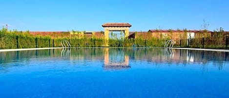 Una piscina azul cristalina con un pabellón en el centro, rodeada de follaje y edificios.