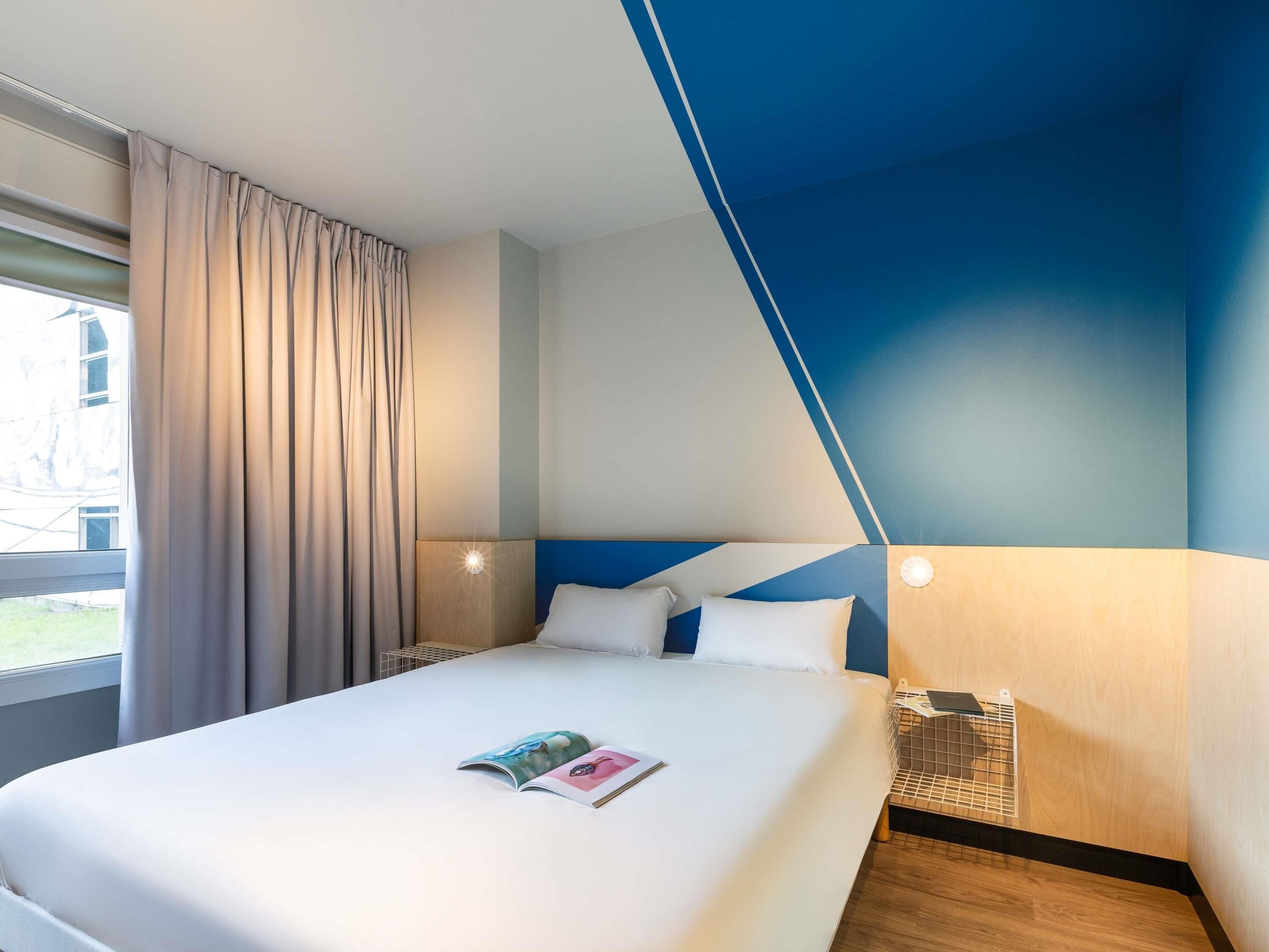Une chambre d’hôtel avec un lit, une table de chevet, une lampe et une fenêtre avec des rideaux.