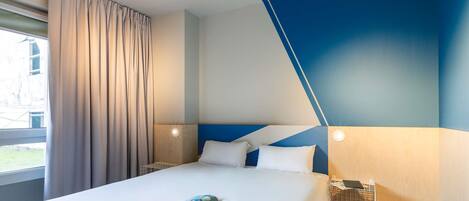 A hotel room with a bed, a nightstand, a lamp, and a window with curtains.